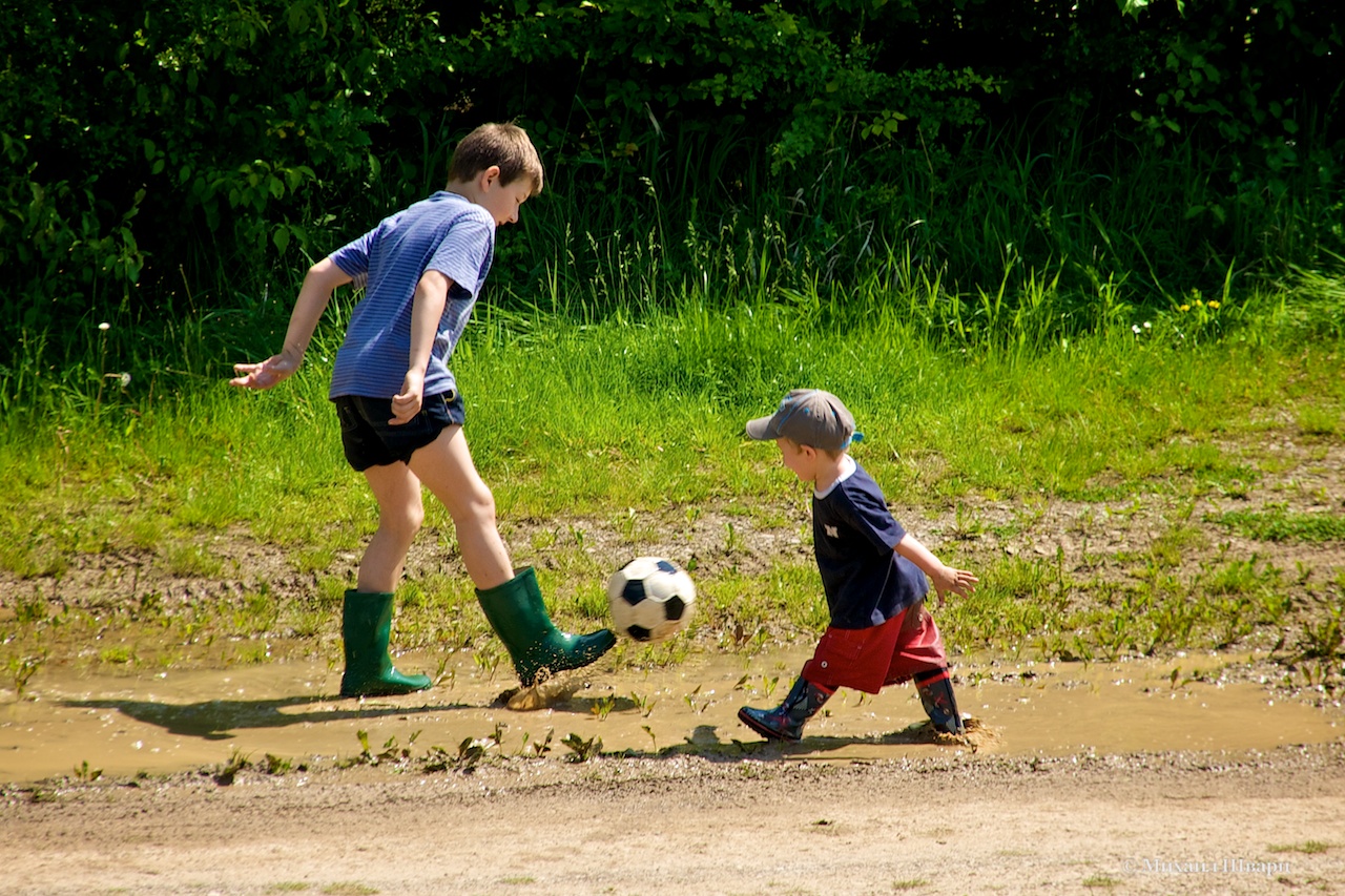 Дети в резиновых сапогах пинают мячик в луже